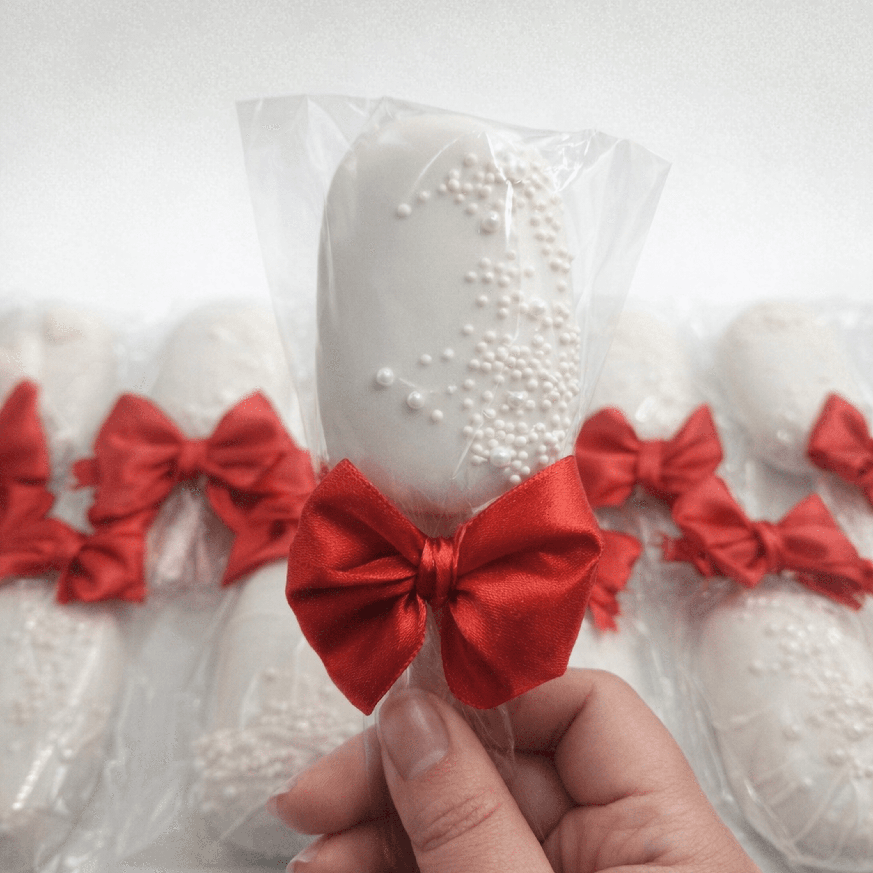 Hand holding a white cakesicle with pearl sprinkles and a red bow in clear packaging.